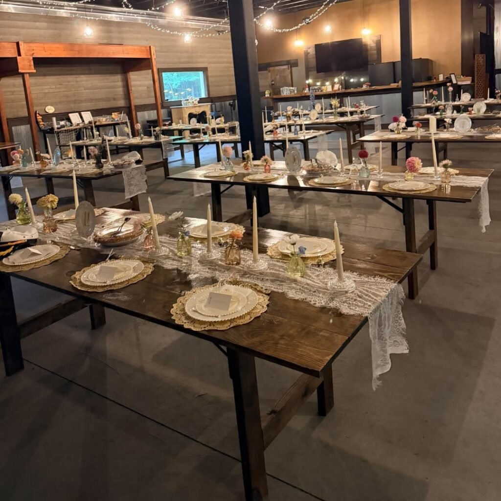 Wooden farmhouse-style tables arranged for a rustic indoor wedding reception with warm lighting.