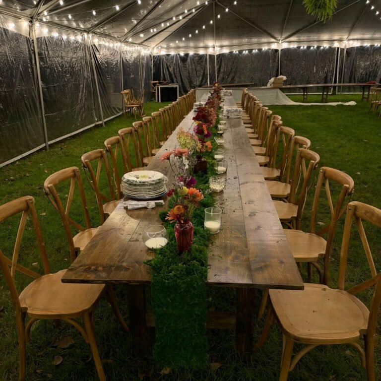 Rustic farmhouse wedding tables decorated with floral centerpieces beneath a lighted outdoor wedding tent.